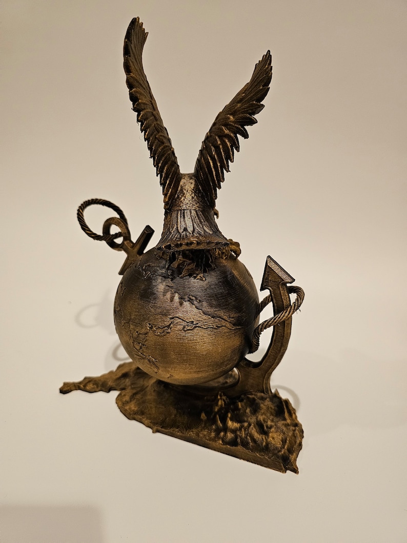 May include: A gold-colored eagle sculpture with its wings spread wide, perched atop a globe with a rope-wrapped anchor. The globe is resting on a textured base.