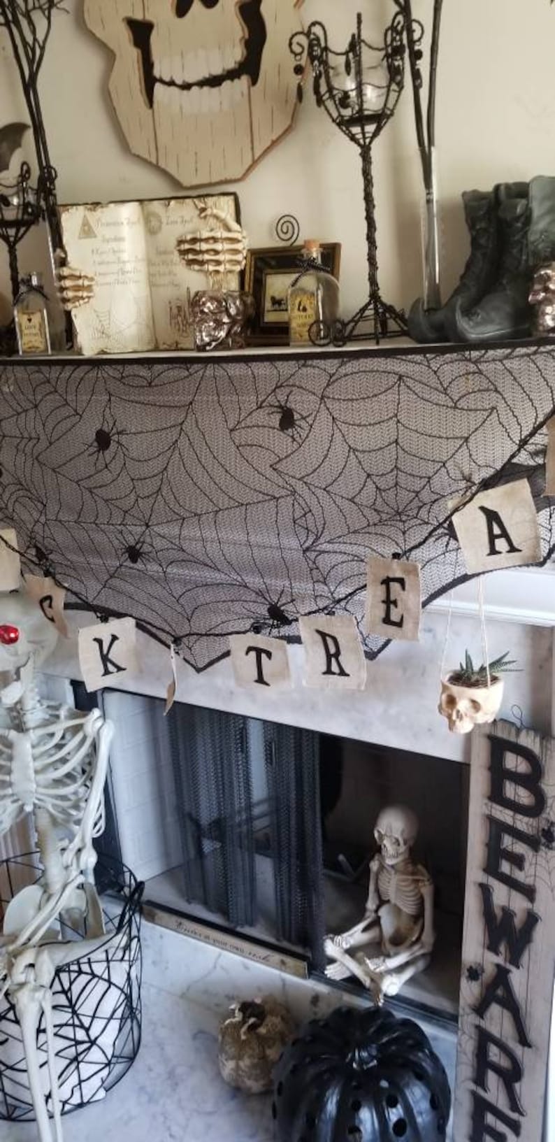 May include: A black and white spiderweb garland with the word "TREAT" written on it hangs above a fireplace. A small succulent plant is in a skull planter. Two skeletons are sitting on the hearth. A black pumpkin with holes in it is on the floor. A sign that says "BEWARE" is on the right side of the fireplace.