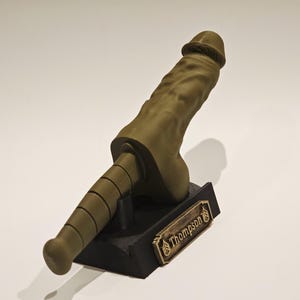 May include: A 3D-printed sculpture of a phallus in a muted olive green color. The sculpture is mounted on a black base with a gold-colored nameplate that reads "Thompson". The sculpture is approximately 10 inches tall.