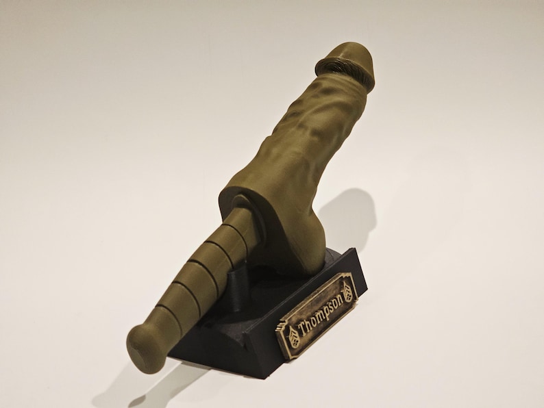 May include: A 3D-printed sculpture of a phallus in a muted olive green color. The sculpture is mounted on a black base with a gold-colored nameplate that reads "Thompson". The sculpture is approximately 10 inches tall.