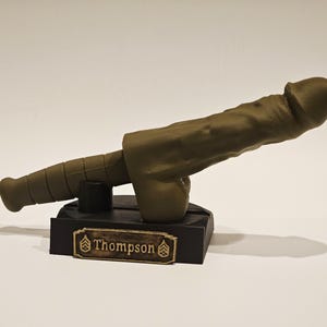 May include: A novelty item resembling a cannon, cast in a muted olive green hue. The cannon is mounted on a black base with a gold-colored nameplate that reads "Thompson". The design is detailed and stylized.
