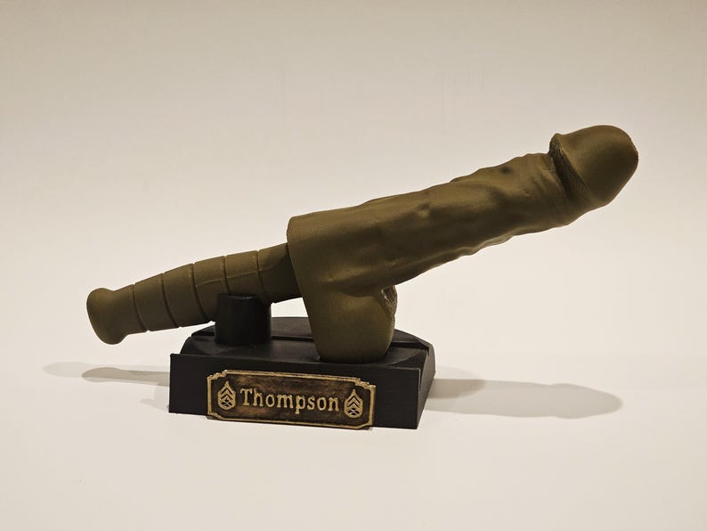 May include: A novelty item resembling a cannon, cast in a muted olive green hue. The cannon is mounted on a black base with a gold-colored nameplate that reads "Thompson". The design is detailed and stylized.