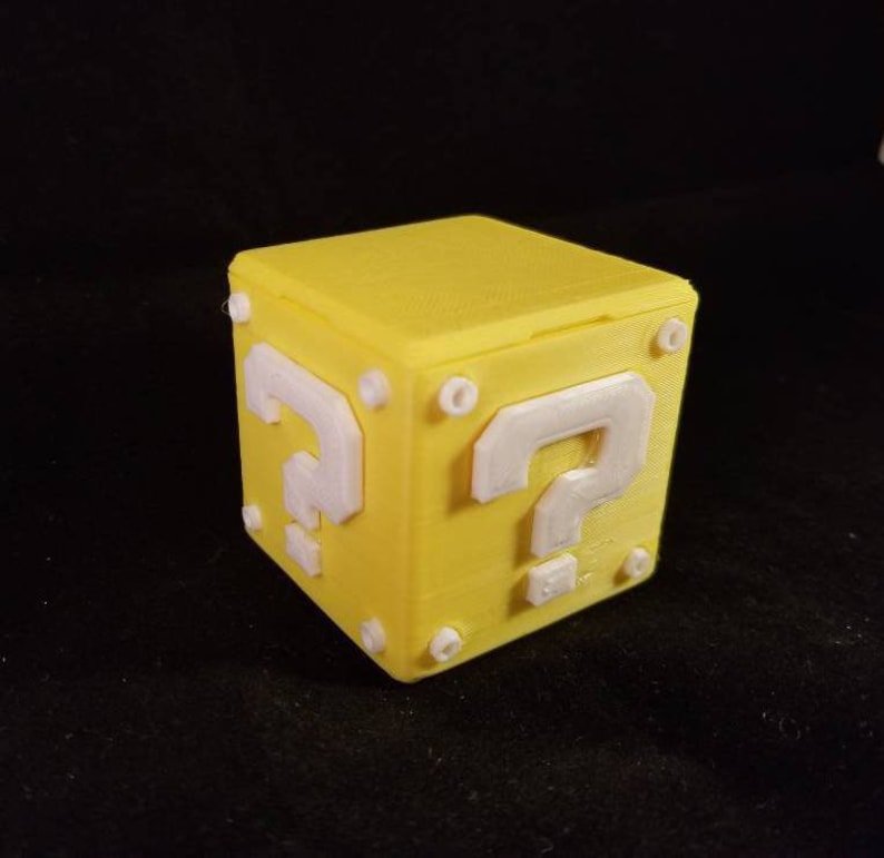 K&ouml;nnte beinhalten: Ein gelber, 3D-gedruckter Fragezeichenblock mit einem wei&szlig;en Fragezeichen auf jeder Seite. Der Block ist ein beliebtes Element in der Super Mario Bros. Videospielserie.