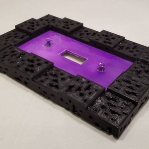 May include: A rectangular, black, textured frame with a purple interior. The frame has a series of cut-out patterns and two silver screws. The center has a rectangular opening.
