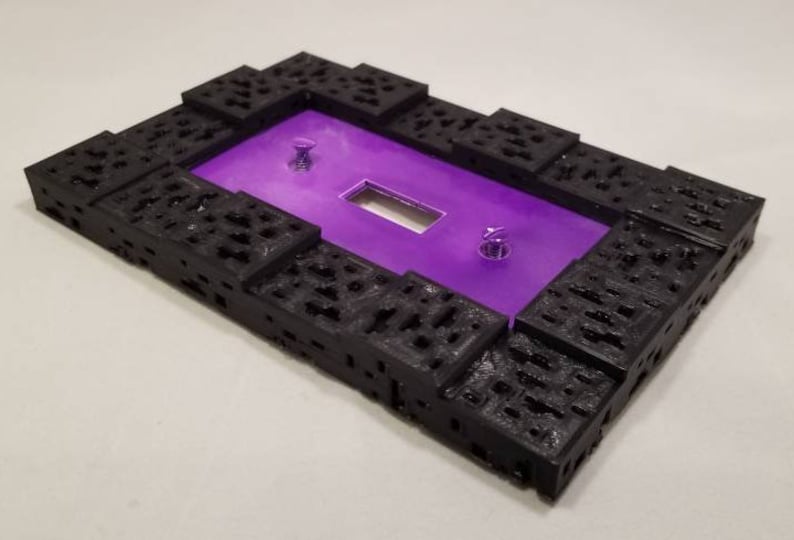 May include: A rectangular, black, textured frame with a purple interior. The frame has a series of cut-out patterns and two silver screws. The center has a rectangular opening.