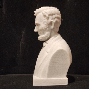 May include: A white bust sculpture of a man with a beard and short hair. The sculpture is on a white base.