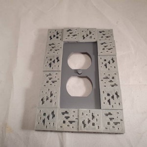 Peut inclure: Une plaque de prise de courant grise avec une bordure d&eacute;corative de petits carr&eacute;s avec un motif pix&eacute;lis&eacute;.