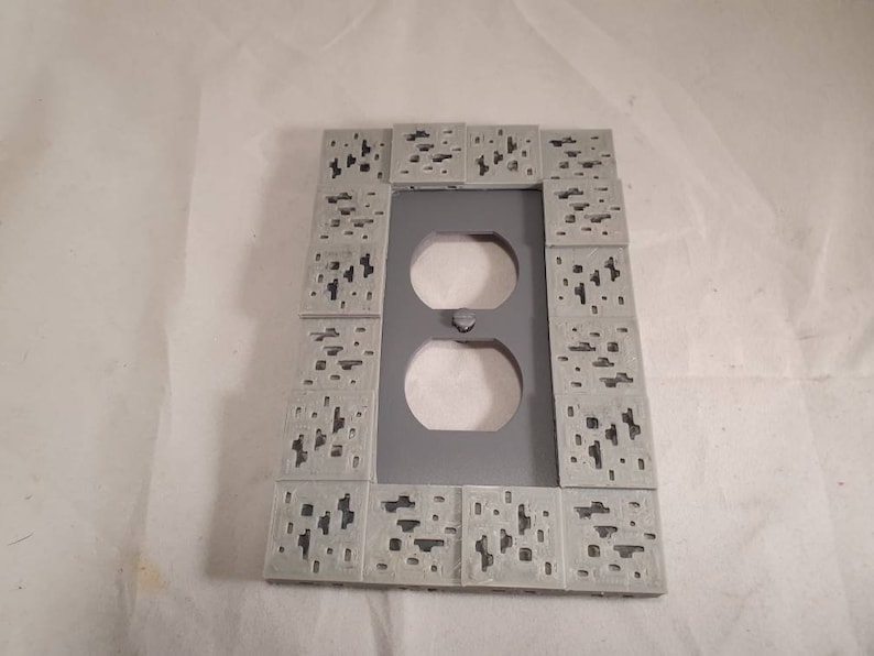 Peut inclure: Une plaque de prise de courant grise avec une bordure d&eacute;corative de petits carr&eacute;s avec un motif pix&eacute;lis&eacute;.