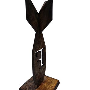 May include: A dark brown, wooden sculpture of a stylized bomb shape with a square base. The letter "F" is cut out of the bomb's body. The sculpture has a rustic, weathered appearance.