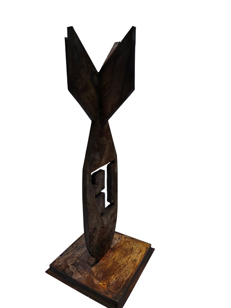 May include: A dark brown, wooden sculpture of a stylized bomb shape with a square base. The letter "F" is cut out of the bomb's body. The sculpture has a rustic, weathered appearance.