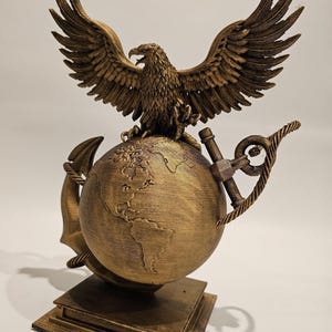 May include: A bronze-colored sculpture featuring an eagle with outstretched wings atop a globe. An anchor and rope are wrapped around the globe. The sculpture is mounted on a square base. The eagle's wings are detailed, and the globe depicts continents.