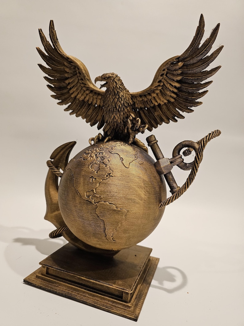 May include: A bronze-colored sculpture featuring an eagle with outstretched wings atop a globe. An anchor and rope are wrapped around the globe. The sculpture is mounted on a square base. The eagle's wings are detailed, and the globe depicts continents.