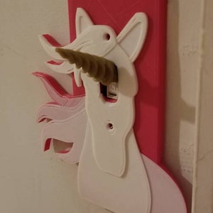 May include: A pink and white 3D printed light switch cover shaped like a unicorn with a gold horn. The horn is sticking out of the light switch.