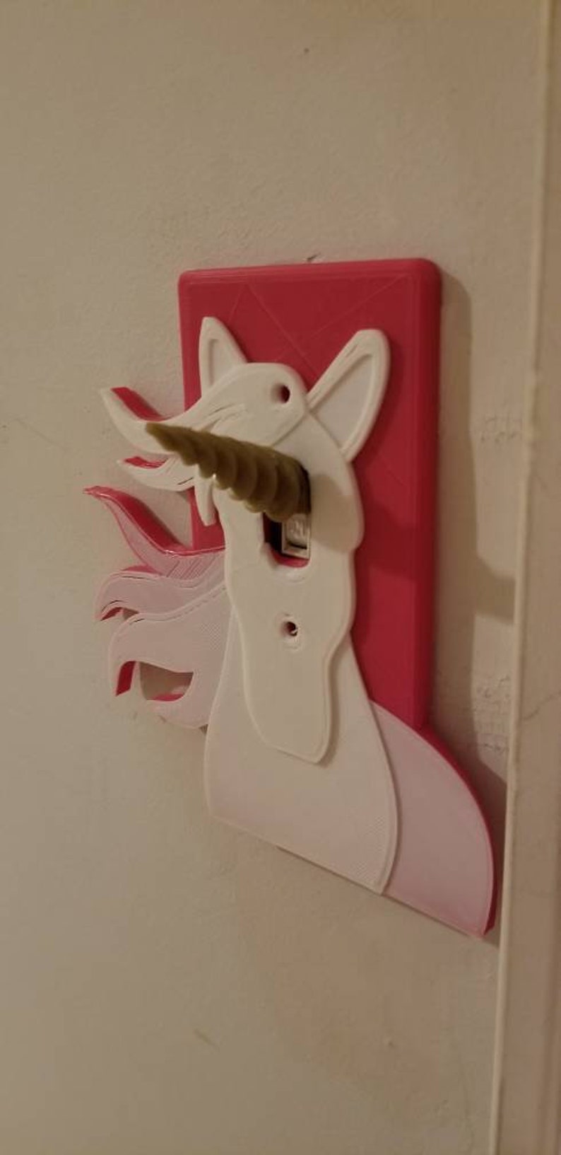 May include: A pink and white 3D printed light switch cover shaped like a unicorn with a gold horn. The horn is sticking out of the light switch.