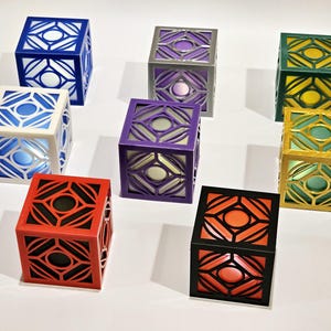 K&ouml;nnte beinhalten: Acht dekorative w&uuml;rfelf&ouml;rmige Boxen mit einem geometrischen Muster und einem Licht im Inneren. Die Boxen sind in verschiedenen Farben, darunter Blau, Wei&szlig;, Lila, Grau, Gr&uuml;n, Gold, Rot und Schwarz.