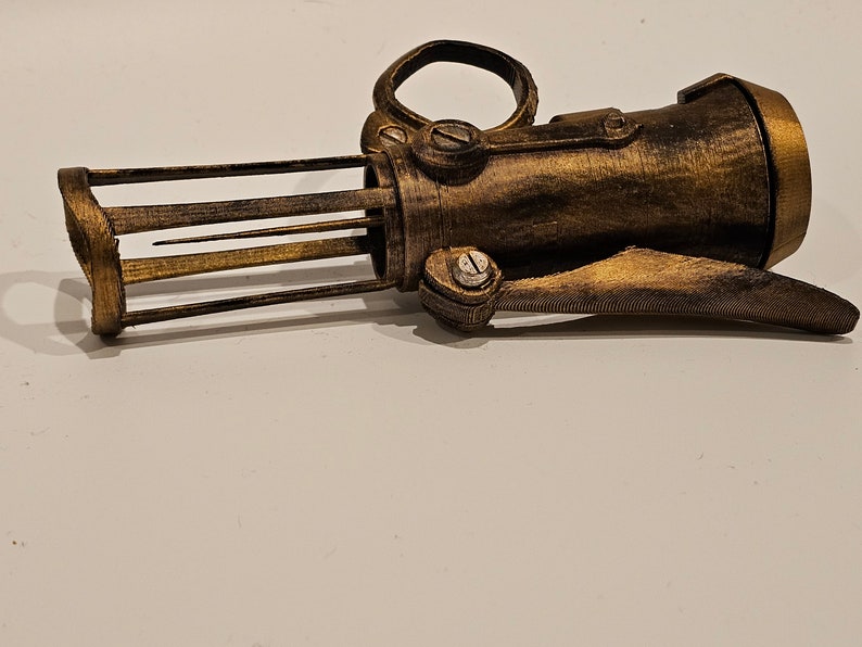 May include: A gold-colored, 3D printed, steampunk-style hand-held weapon with a cage-like front and a lever on the side.
