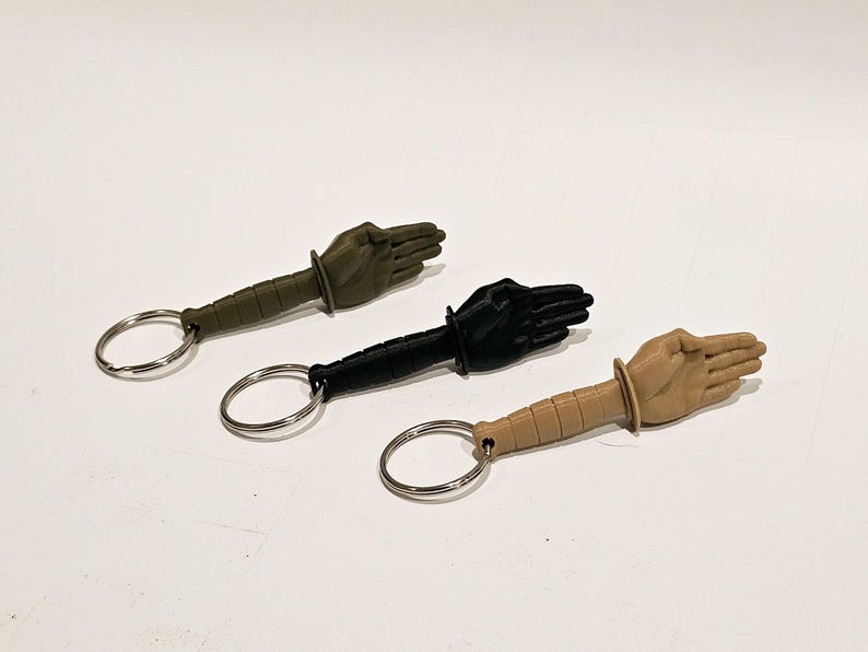 May include: Three keychains with a hand-shaped design. The keychains are green, black, and tan. Each keychain has a metal ring.