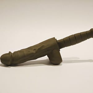 May include: A novelty item in the shape of a phallus and a knife handle. The item is olive green and appears to be made of a solid material. The handle has a ridged design. The item is set against a white background.