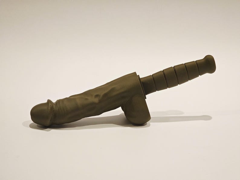 May include: A novelty item in the shape of a phallus and a knife handle. The item is olive green and appears to be made of a solid material. The handle has a ridged design. The item is set against a white background.