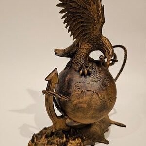 May include: A gold-colored eagle sculpture with outstretched wings perched on a globe, anchored by a rope-wrapped anchor. The globe has a detailed map of the world.