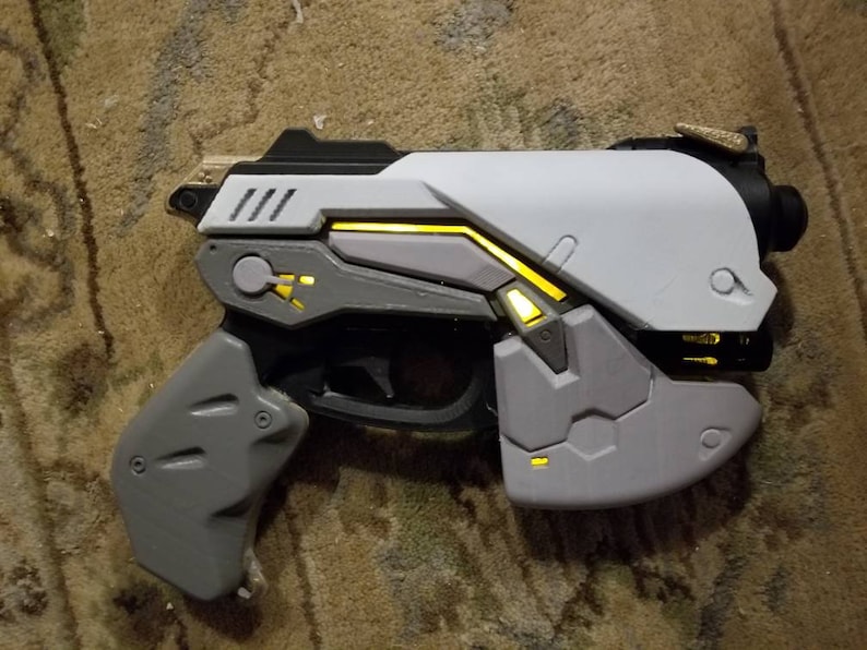Puede incluir: Una pistola de juguete de pl&aacute;stico blanco y gris con detalles amarillos. La pistola tiene un agarre texturizado y un dise&ntilde;o futurista.