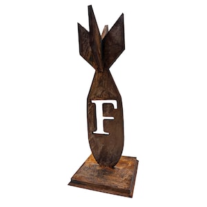 May include: A rustic metal sculpture of a bomb shape with the letter "F" cut out. The sculpture has a brown, weathered finish and sits on a square base. The fins at the top are pointed.