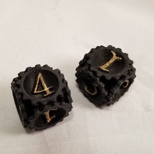 May include: Two black dice with gold numbers. The dice are shaped like gears and have a textured surface. One die shows the number 4 and the other die shows the number 1.