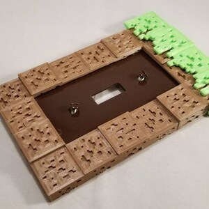 May include: A light brown and green 3D printed light switch cover with a Minecraft-inspired design. The cover is made to look like a block of dirt with grass on top.
