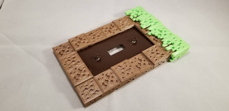 May include: A light brown and green 3D printed light switch cover with a Minecraft-inspired design. The cover is made to look like a block of dirt with grass on top.
