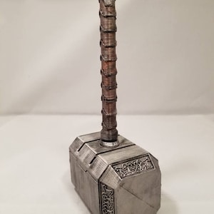 Op de afbeelding: Een zilverkleurige Mjolnir-hamer met een bruine houten handvat. De hamerkop heeft ingewikkelde details en een getextureerd oppervlak.