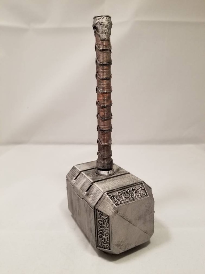 Op de afbeelding: Een zilverkleurige Mjolnir-hamer met een bruine houten handvat. De hamerkop heeft ingewikkelde details en een getextureerd oppervlak.
