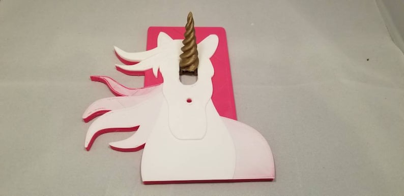 May include: A pink and white unicorn light switch cover with a gold horn. The unicorn's mane is flowing in the wind.