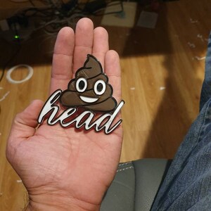 May include: A brown 3D printed poop emoji with a white smiling face and the word "head" printed in white below it.