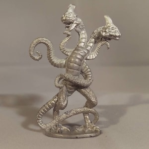 May include: A silver-colored metal figurine of a three-headed creature with a serpentine body. The creature has a long, slender neck and a small head with a pointed snout. The figurine is standing on a small base.