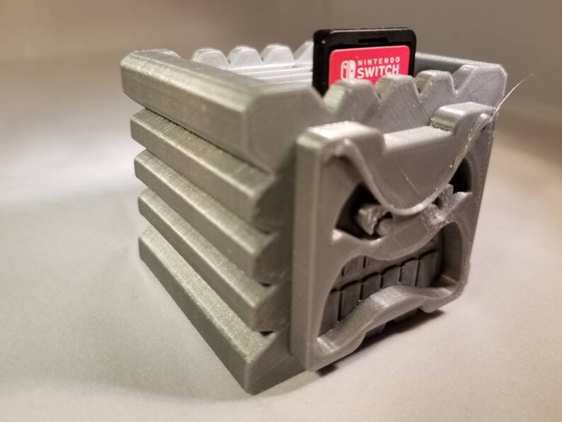 K&ouml;nnte beinhalten: Ein 3D-gedruckter grauer Spielkartuschenhalter in Form eines Gesichts mit einem missbilligenden Ausdruck. Der Halter hat einen Schlitz f&uuml;r eine Nintendo Switch-Spielkartusche. Die Kartusche ist mit "Nintendo Switch" beschriftet.