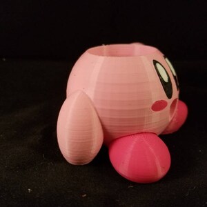 May include: A pink 3D printed planter shaped like the video game character Kirby. The planter has a hole in the top for planting and a small hole on the side for drainage.
