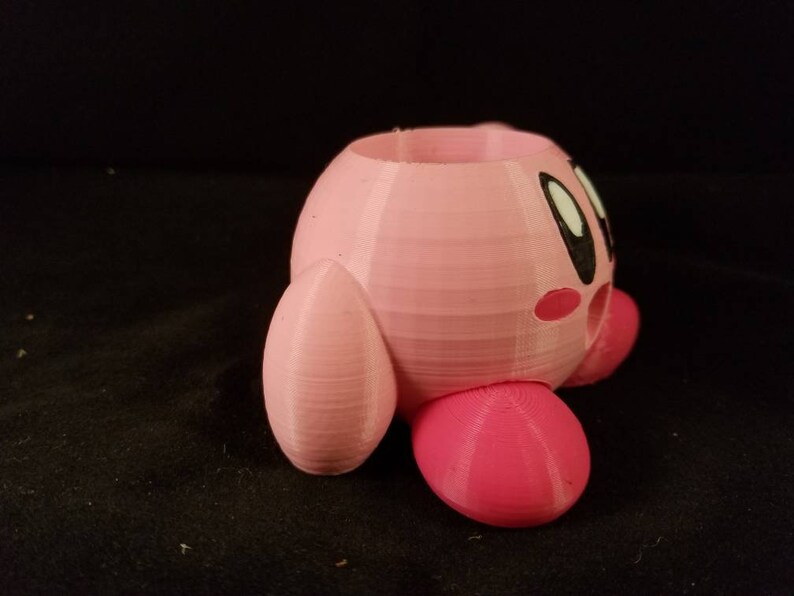 May include: A pink 3D printed planter shaped like the video game character Kirby. The planter has a hole in the top for planting and a small hole on the side for drainage.