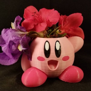 May include: A pink 3D printed planter shaped like a cartoon character with large eyes and a wide smile. The planter is filled with purple irises and red flowers.