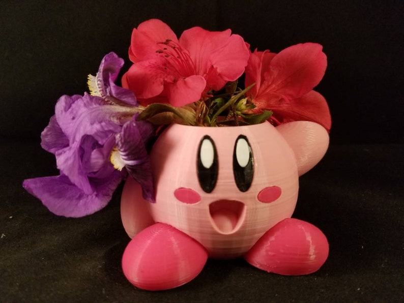 May include: A pink 3D printed planter shaped like a cartoon character with large eyes and a wide smile. The planter is filled with purple irises and red flowers.