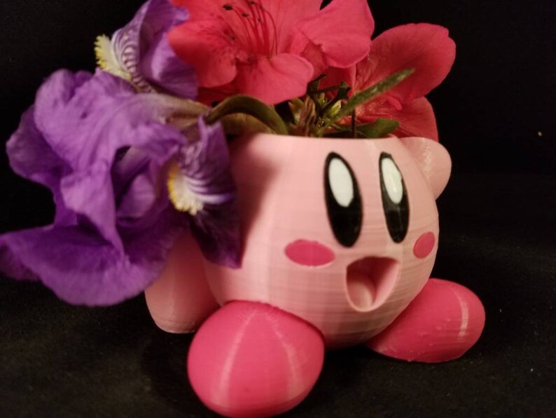 May include: A pink 3D printed planter shaped like the video game character Kirby, with a purple iris flower and red azalea flowers inside.