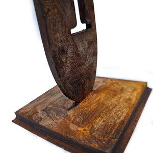 May include: A rustic wooden sculpture featuring a stylized letter 'I' cut out of a dark brown, textured piece. The sculpture is mounted on a rectangular base, also made of wood, with a similar aged appearance. The overall design has a natural, handcrafted feel.
