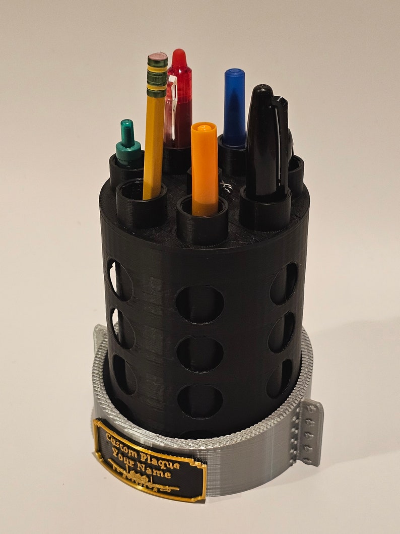 May include: Black plastic pencil holder with a gray base and a gold plaque that says "Custom Plaque Your Name". The holder has multiple slots for pens and pencils. There are six writing utensils in the holder, including a red pen, a blue pen, a yellow highlighter, a green pen, a black pen, and a yellow pencil.