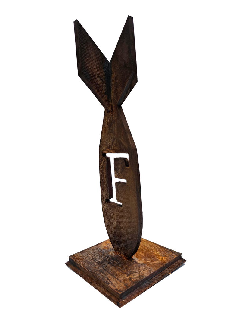 May include: A brown, stylized metal sculpture of a bomb shape with the letter "F" cut out. The sculpture has a textured finish and is mounted on a square base. The sculpture is a decorative item.