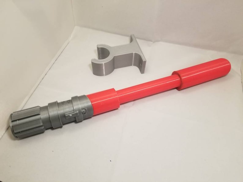 May include: A red and gray lightsaber prop with a matching gray stand. The lightsaber features a red cylindrical handle and a gray detailed hilt. The stand is a simple gray shape.