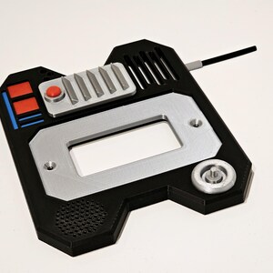 May include: A black and silver 3D printed device with a red button, a rectangular opening, and a small round knob. The device has a textured surface and a small antenna.