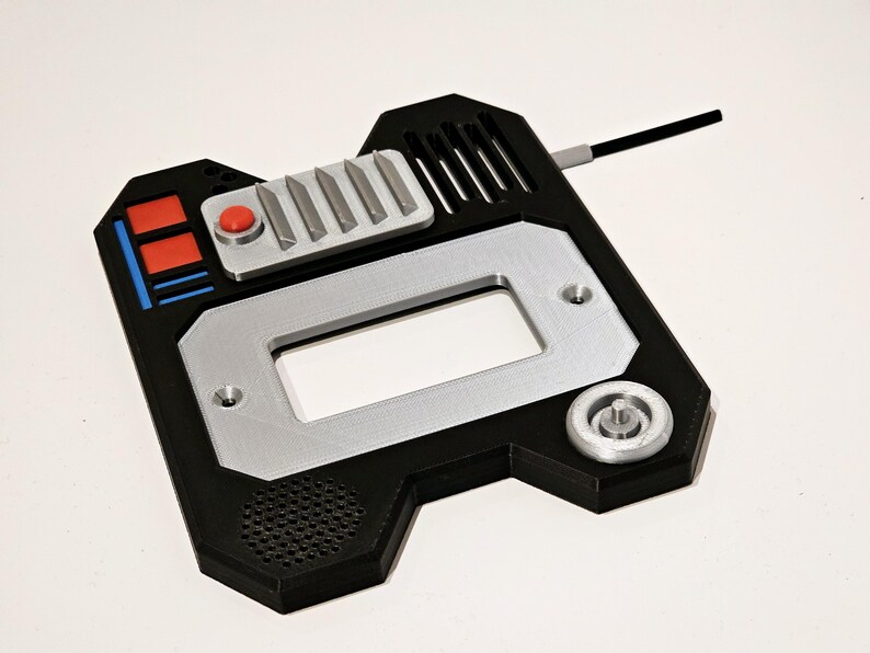 May include: A black and silver 3D printed device with a red button, a rectangular opening, and a small round knob. The device has a textured surface and a small antenna.