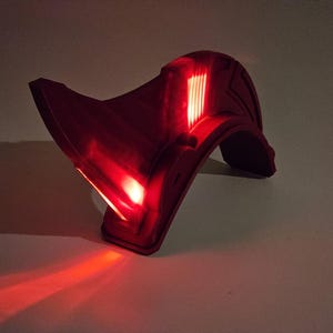 Op de afbeelding: Een rode plastic helm met een verlicht vizier. Het vizier heeft een reeks rode lampjes die aanstaan.