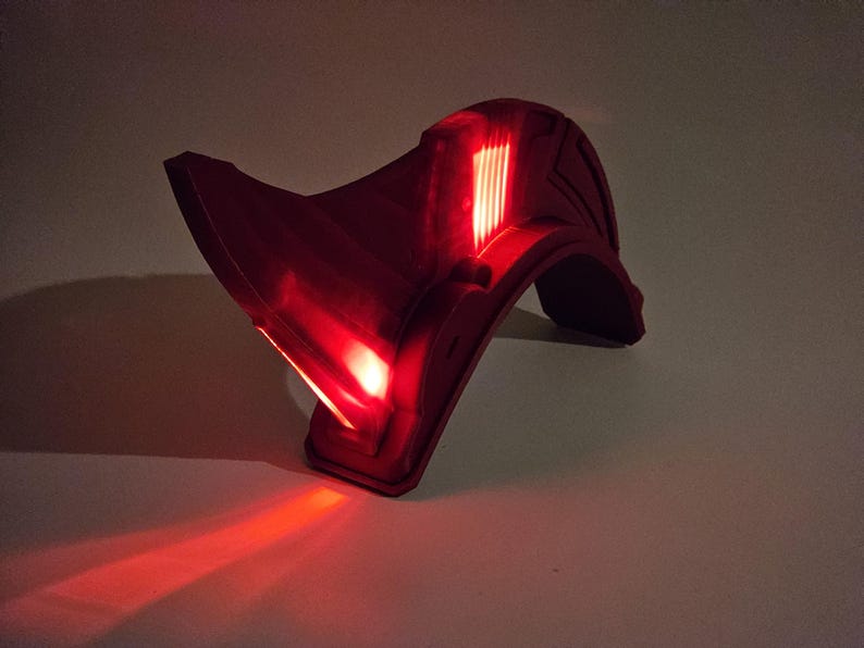Op de afbeelding: Een rode plastic helm met een verlicht vizier. Het vizier heeft een reeks rode lampjes die aanstaan.