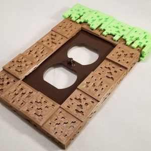 May include: A brown and green 3D printed outlet cover with a grass block design. The cover is shaped like a Minecraft block.