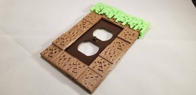 May include: A brown and green 3D printed outlet cover with a grass block design. The cover is shaped like a Minecraft block.